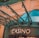 The image displays the entrance to a casino, characterized by detailed and ornate architectural elements with green metal structures and a sign that reads 'CASINO'. The background includes a glass dome and a peach-colored wall.