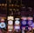 A lively casino scene featuring a row of bright, colorful slot machines with themes like Monopoly, Michael Jackson, and Lord of the Rings. The gaming machines display a variety of flashing lights and digital screens, creating an energetic and exciting atmosphere. Reflections of the lights are visible in glossy surfaces above the machines.