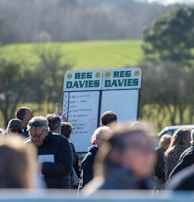 A group of people gathered outdoors with two large betting boards in the background labeled 'REG DAVIES'. The setting appears to be a countryside location with trees and fields visible in the distance. The crowd consists of individuals dressed in jackets and hats, engaging in conversation.